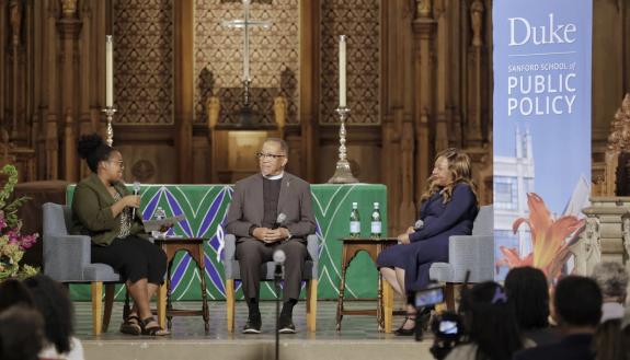 Three people sit in the front of the Duke Chapel next to a banner that reads, "Duke Sanford School of Public Policy"