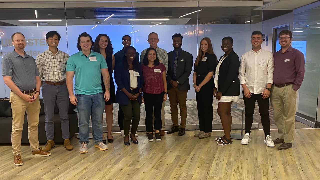 13 people stand, smiling, in a line facing the camera in a classroom in the Duke in DC office.