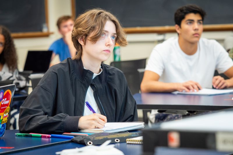 Meehan sitting at a desk in a classroom, wearing a black jacket, writing in a notebook with a purple pen