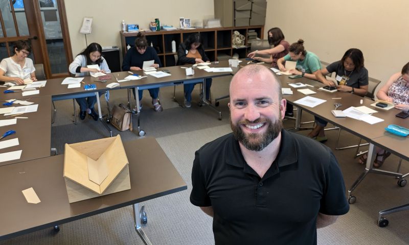 A man stands and smiles in front of classroom of bookbinding workshop students